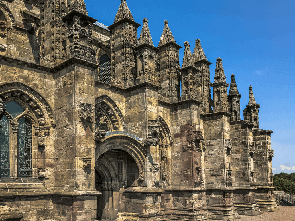 Exterior view of Rosslyn Chapel in Scotland, showcasing intricate Gothic stonework and spires under a clear blue sky.