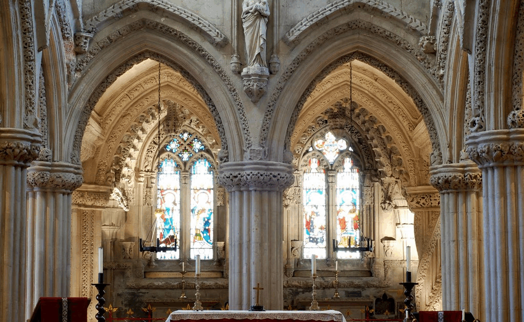 Interior view of Rosslyn Chapel showing the altar, ribbed arches, and stained glass windows illuminated by natural light.