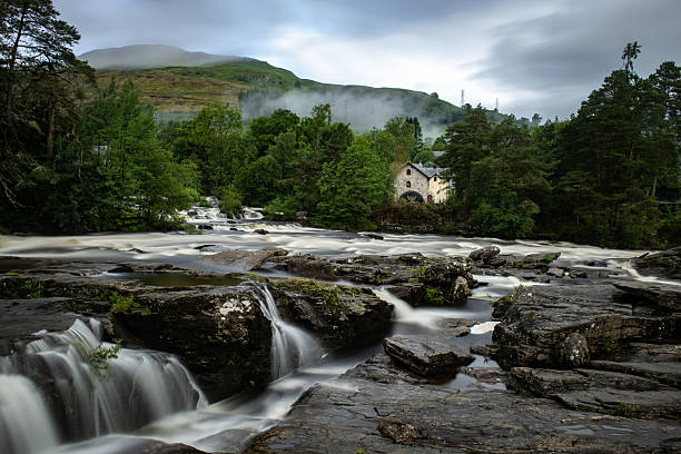 Water rushing over the rocky Falls of Dochart surrounded by forested hills and a misty Highland backdrop